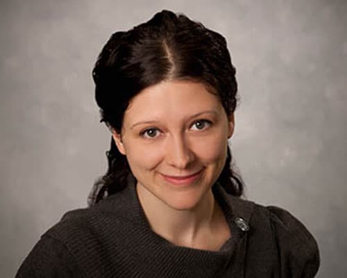 Person with long dark hair and a neutral expression, wearing a dark top, against a gray backdrop.