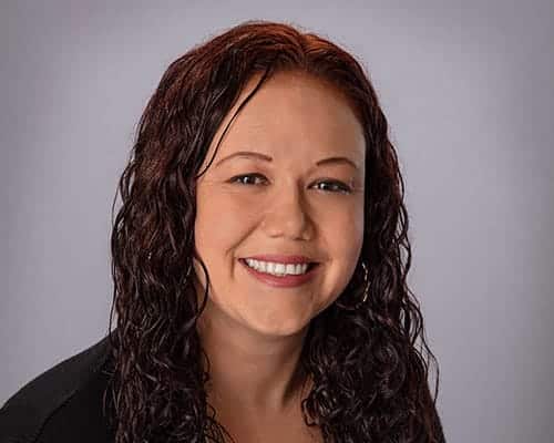 A person with long, curly brown hair smiles at the camera against a plain gray background.