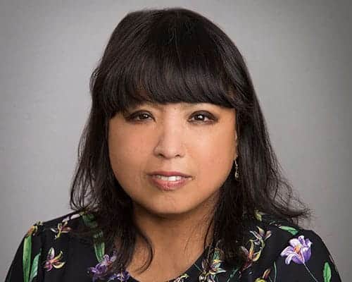 A person with long dark hair wearing a floral-patterned top, looking directly at the camera with a neutral expression.