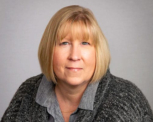 A person with shoulder-length blonde hair is wearing a gray sweater over a collared shirt against a plain background.