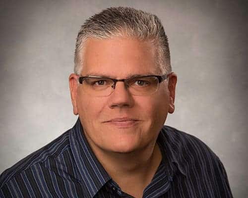 A person with short gray hair, glasses, and a striped shirt looks directly at the camera against a neutral background.