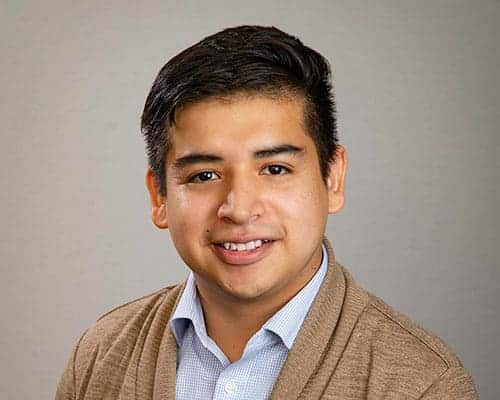 Portrait of a person with short dark hair, wearing a blue shirt and a brown cardigan, set against a neutral background.