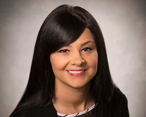 A woman with long dark hair and a black outfit smiles against a gray background.