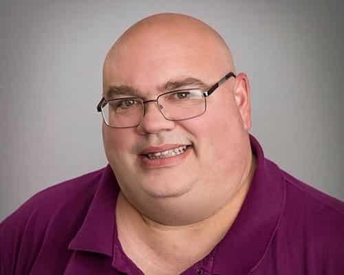 A person with glasses and a bald head is wearing a magenta shirt and smiling at the camera.