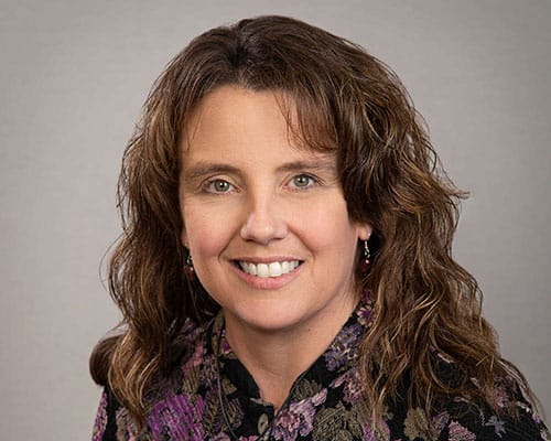 A woman with curly brown hair smiles at the camera, wearing a floral top, against a gray background.