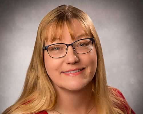 A person with long blonde hair and glasses smiles at the camera against a gray background.