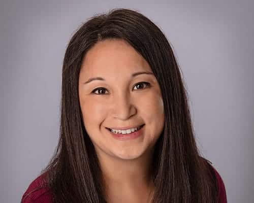 Portrait of a person with long dark hair, smiling at the camera, wearing a burgundy top against a plain gray background.