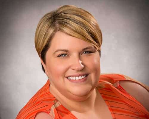 A person with short hair smiles, wearing an orange top with stripes, against a gray background.