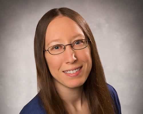 A person with long, straight brown hair and glasses smiles at the camera, wearing a blue top against a neutral background.