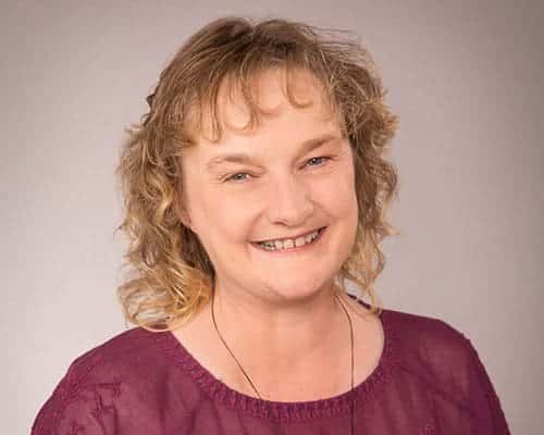 Smiling woman with curly, shoulder-length hair wearing a purple top against a neutral background.