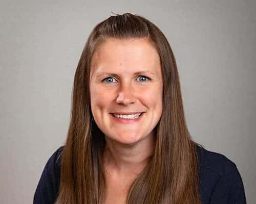 A person with long brown hair smiles against a gray background.