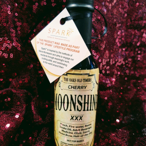 A clear glass bottle filled with homemade moonshine, labeled with a tag that says "Moonshine" on a sparkly cloth.