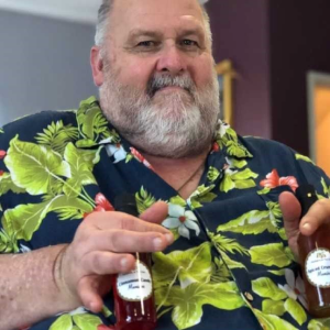 A bearded man in a Hawaiian shirt holds two bottles of moonshine