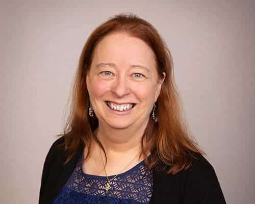 A person with long reddish-brown hair, wearing a blue lace top and black cardigan, smiles against a plain background.