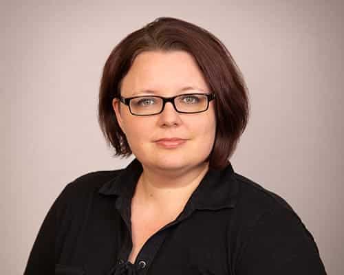 Person with short dark hair and glasses wearing a black top against a neutral background.