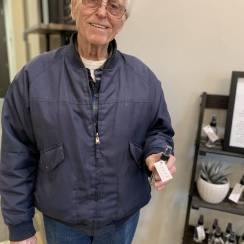 An older man with a kind smile stands confidently in front of a shelf, holding a bottle.