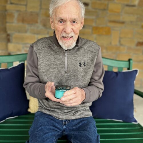 An elderly gentleman enjoying a peaceful moment on a green bench, cradling a refreshing blue cup.