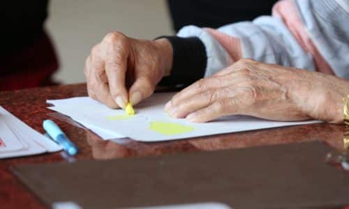 An elderly woman writes on paper with a yellow highlighter, a pen nearby.