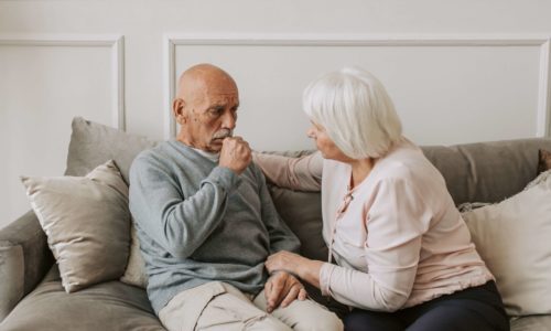 An elderly man sitting on a couch appears thoughtful while an elderly woman gently comforts him.