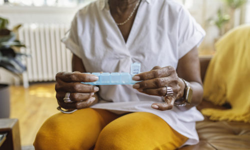 Senior black woman sits on the couch at home and takes medications from a daily pill organizer. Cropped shot does not show the woman's face.