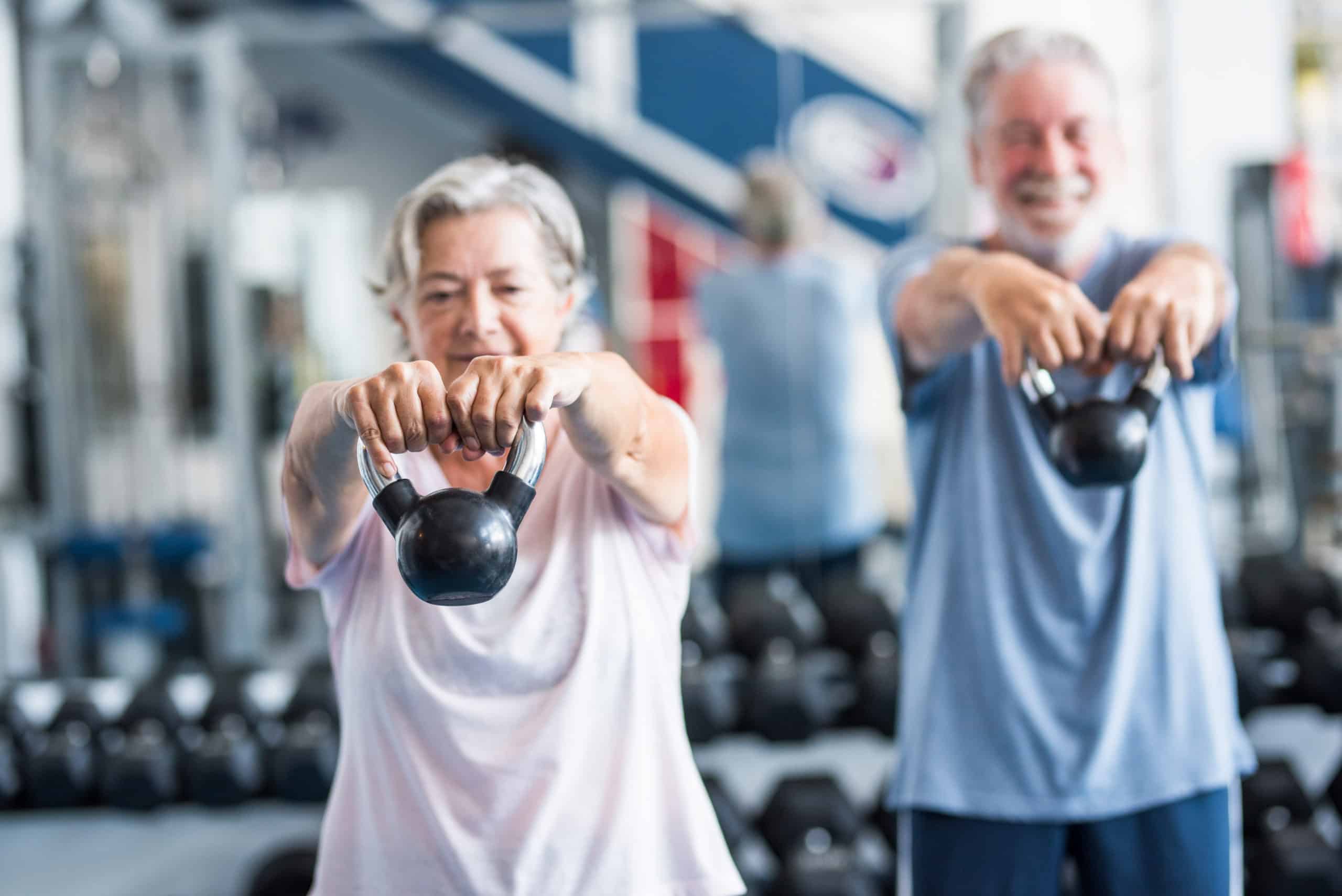 Two older adults lift kettlebells during a workout in a gym.