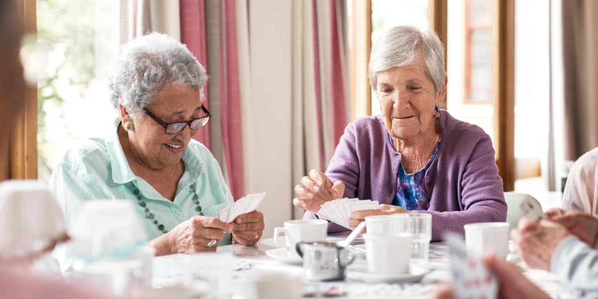 Two elderly women sit at a table playing cards, surrounded by teacups and a coffee pot.
