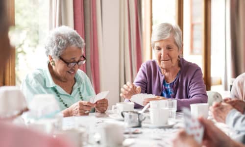 Two elderly women sit at a table playing cards, surrounded by teacups and a coffee pot.