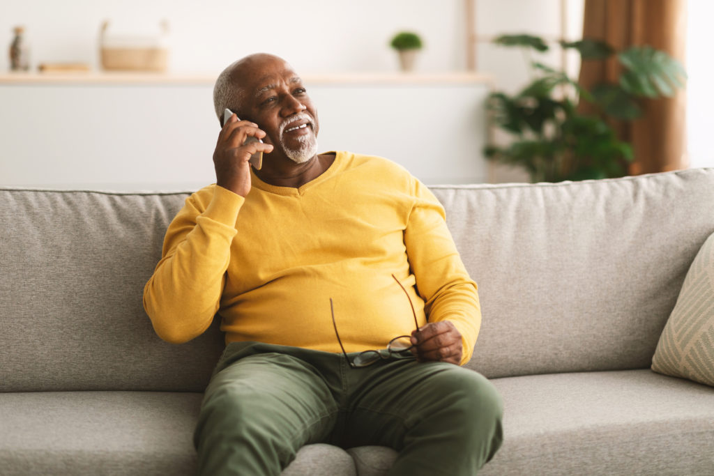 Senior African American Man Talking On Cellphone Having Pleasant Phone Conversation Sitting On Sofa At Home, Looking Aside And Smiling. Mobile Communication Concept
