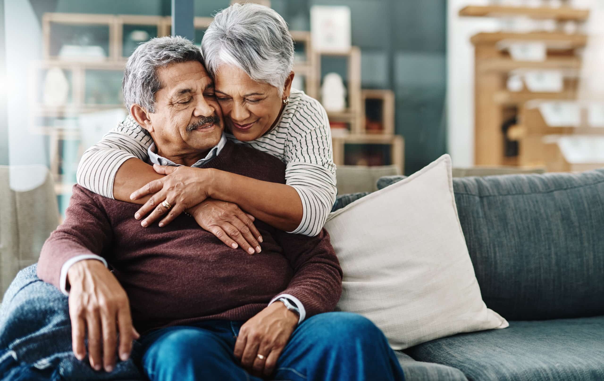 An elderly couple sits on a couch, with the woman embracing the man from behind. They both appear content and relaxed.