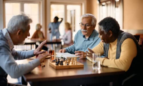 Three elderly men sit around a table playing chess in a social setting, with others in the background.