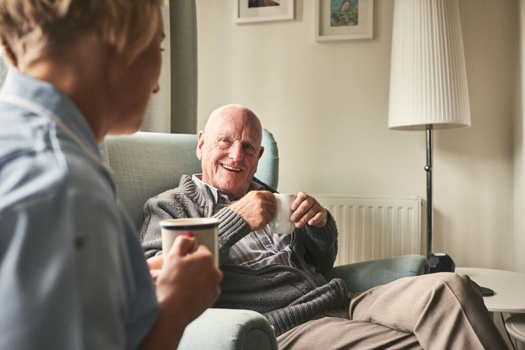 Smiling senior man with cup of coffee talking to female caregiver sitting in front at care home