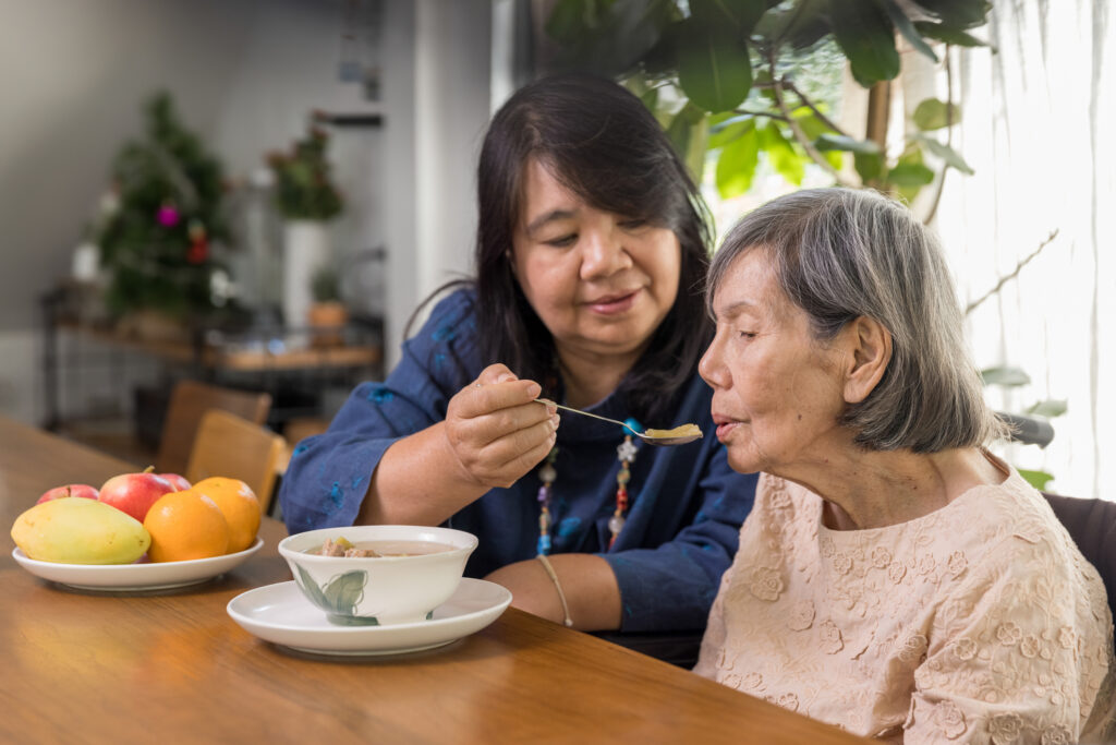 Daughter feeding elderly mother with soup. Senior Asian woman bored with food.