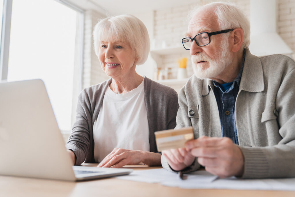 Smart old senior elderly caucasian couple grandparents husband and wife receiving pension paying domestic bills, doing online shopping with credit card and laptop, e-banking and e-commerce at home