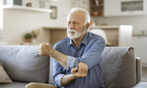 Senior man with Elbow pain.Old male massaging painful hand indoors. Old man hand holding his elbow suffering from elbow pain. Senior man suffering from pain in hand at home. Old age, health