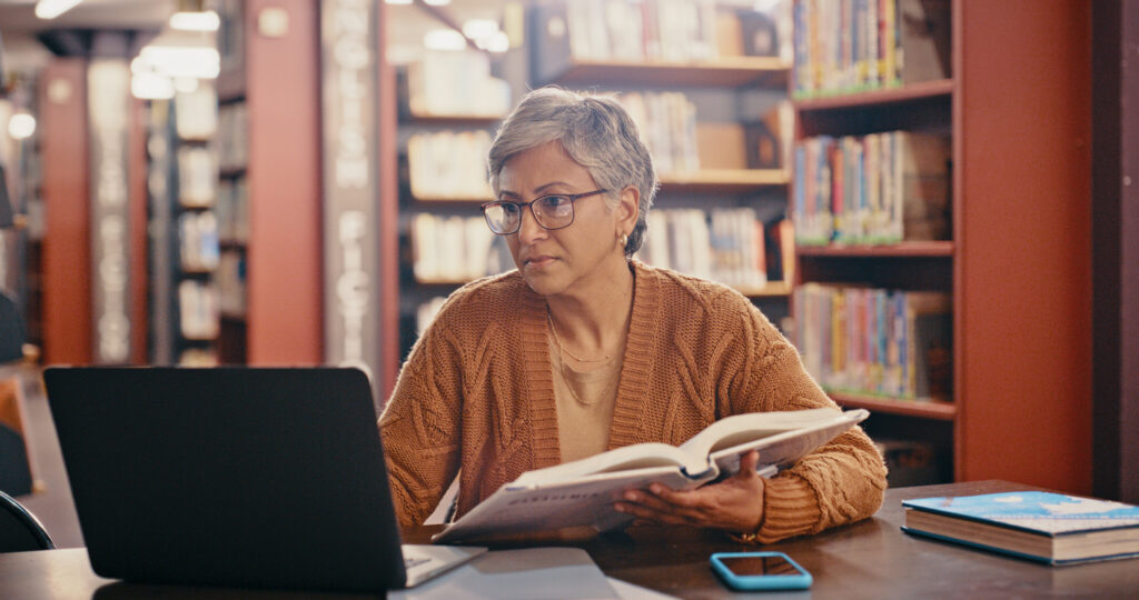 Senior woman, computer and planning in a library with book and laptop for lecture class. Teacher, university professor and education worker reading web data and research for class on a study desk