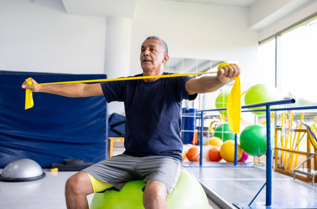 Mature Latin American man doing physiotherapy exercises using a stretch band at a rehabilitation center