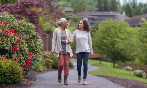 A senior woman links arms with her millennial Eurasian daughter as they happily walk through a natural parkland area and enjoy their time together.