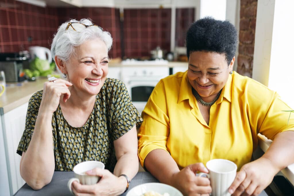 Image of two aged female neighbors drinking coffee together in kitchen.