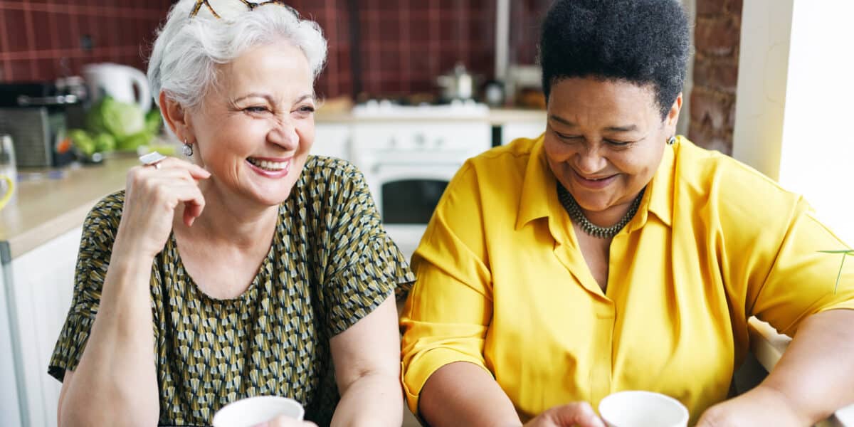 Image of two aged female neighbors drinking coffee together in kitchen.