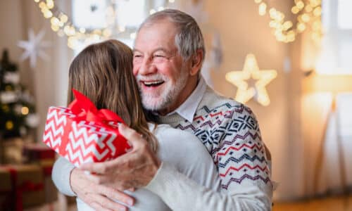 Portrait of young woman giving present to happy grandfather indoors at home at Christmas.