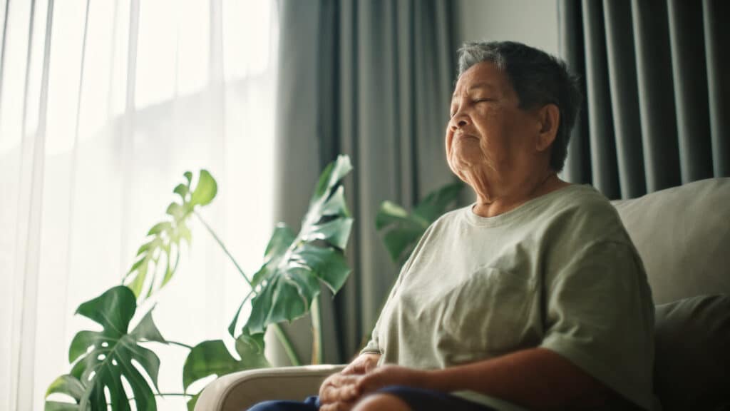 Asian senior woman meditating on sofa at home.